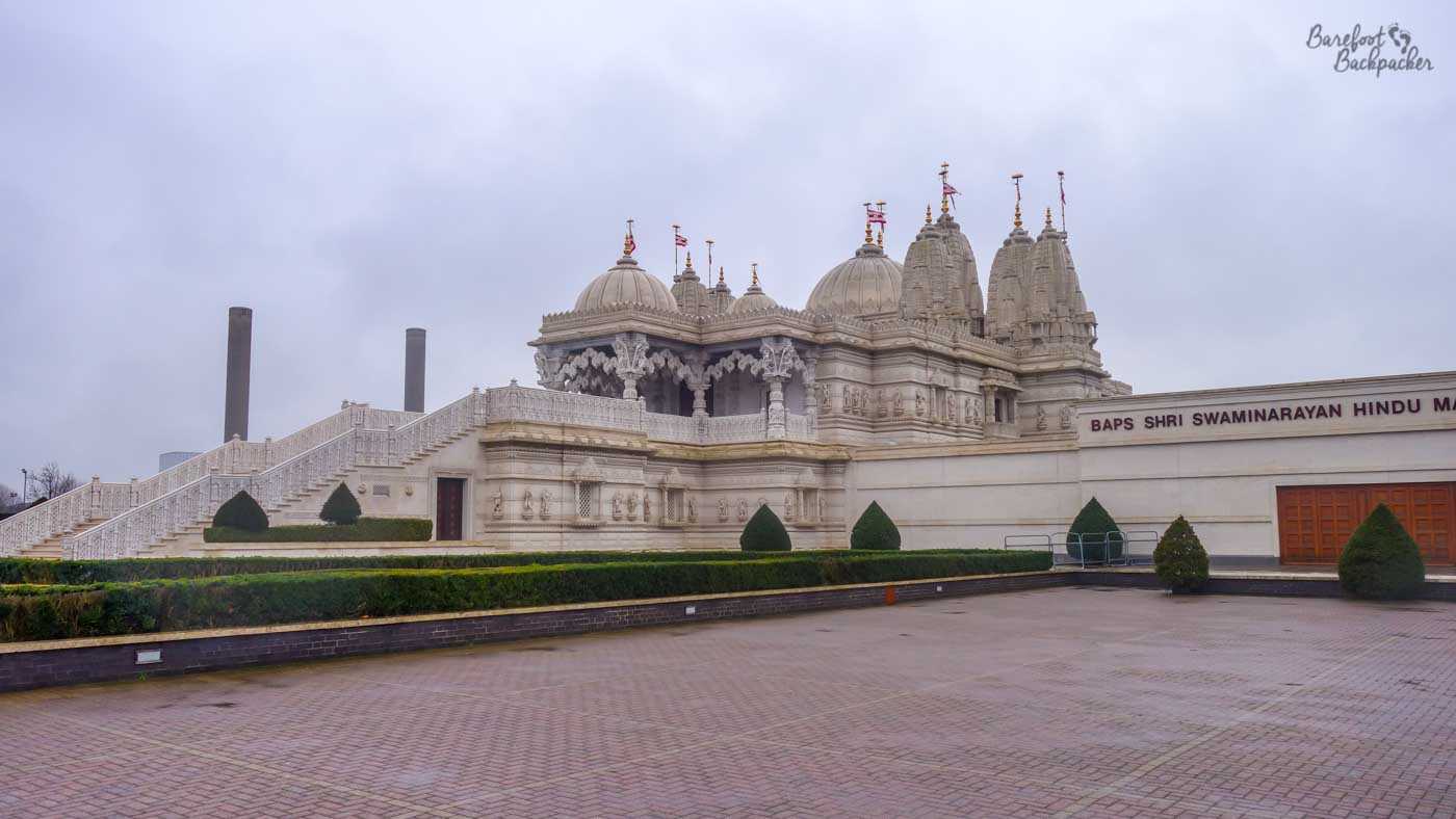 Under grey, cloudy, overcast skies there is a large building. It's set on a raised patio area. It is a Hindu temple. It is almost entirely white, with a large staircase on the left of shot rising gently to a series of ornately carved towers. In the midground is the edge oft he patio, bounded by another white ornately-sculpted wall.