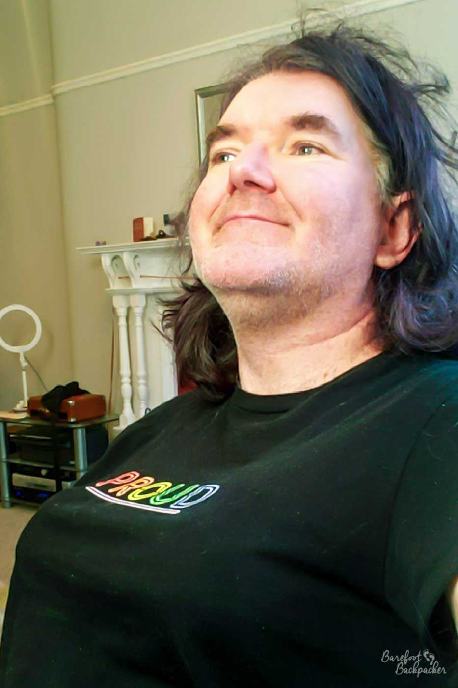 Someone is wearing a black t-shirt with the word 'proud' in rainbow letters on it. They are sat with quite a straight back. Under their t-shirt are what appear to be breasts, although the person in question otherwise presents as a man.