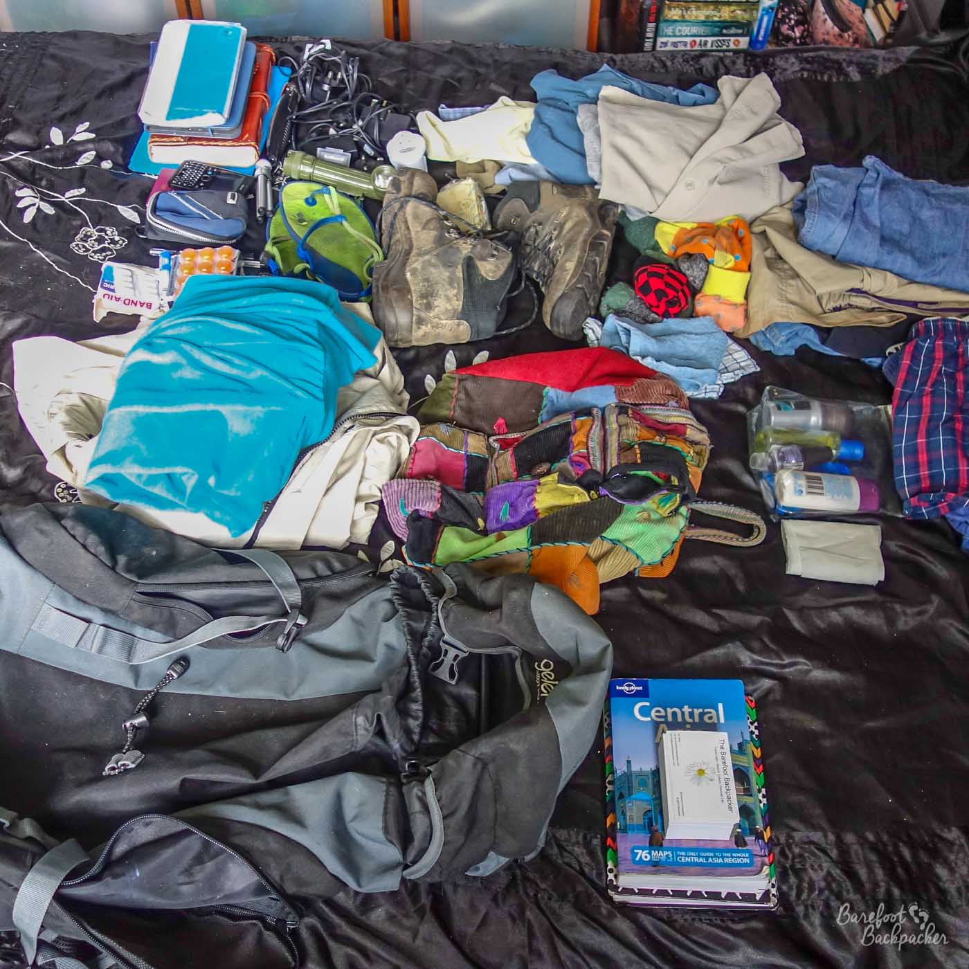 On a bed are lots of items, including separate piles of types of clothing, a pair of walking boots, a backpack, some paper/pads/pens, electrical items, and a guidebook to Central Asia. Probably a couple of packing mistakes here.