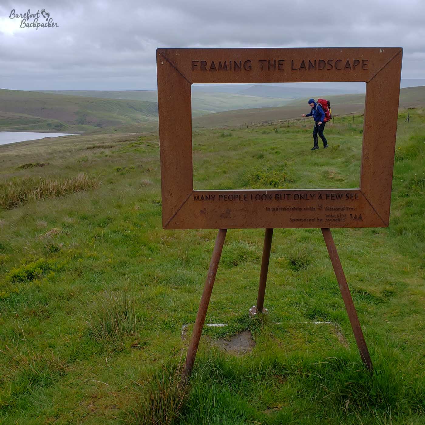 In damp, empty, moorland sloping down toward a reservoir is a large frame, like a picture frame, on three stilts. On the frame is written 'Framing the Landscape. Many people look but only a few see.'. Inside the frame, as it were, stands a backpacker/hiker, standing on the grass behind so their entire body is visible if you look through the frame.