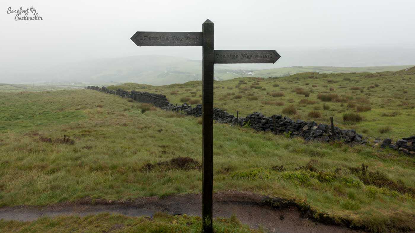 A wooden, dark signpost with two struts – Pennine Way North and Pennine Way South. It stands on grassy, shrubby, damp, moorland in the mist. A dry-stone wall separates two fields into the background.