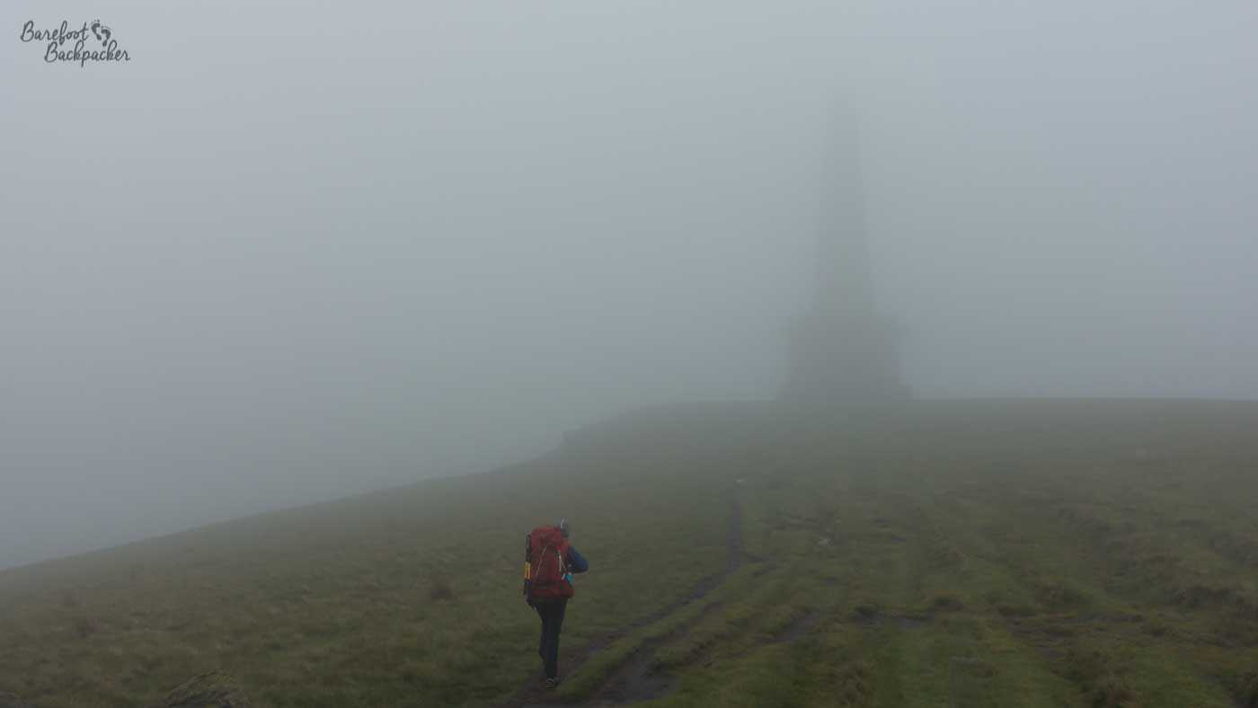 At the bottom of shot is a hiker. The rest of the image is mist/fog. The difference between the land and the sky is gauged by the dark or light level of the mist. Somewhere in the distance is the vague outline of a tower.