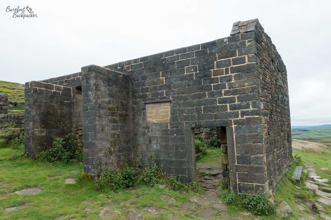 The framework of a dark stone brick building stands on moorland. The basic shape is sound, with a couple of wall juttings suggesting rooms that are lost. There is no roof.