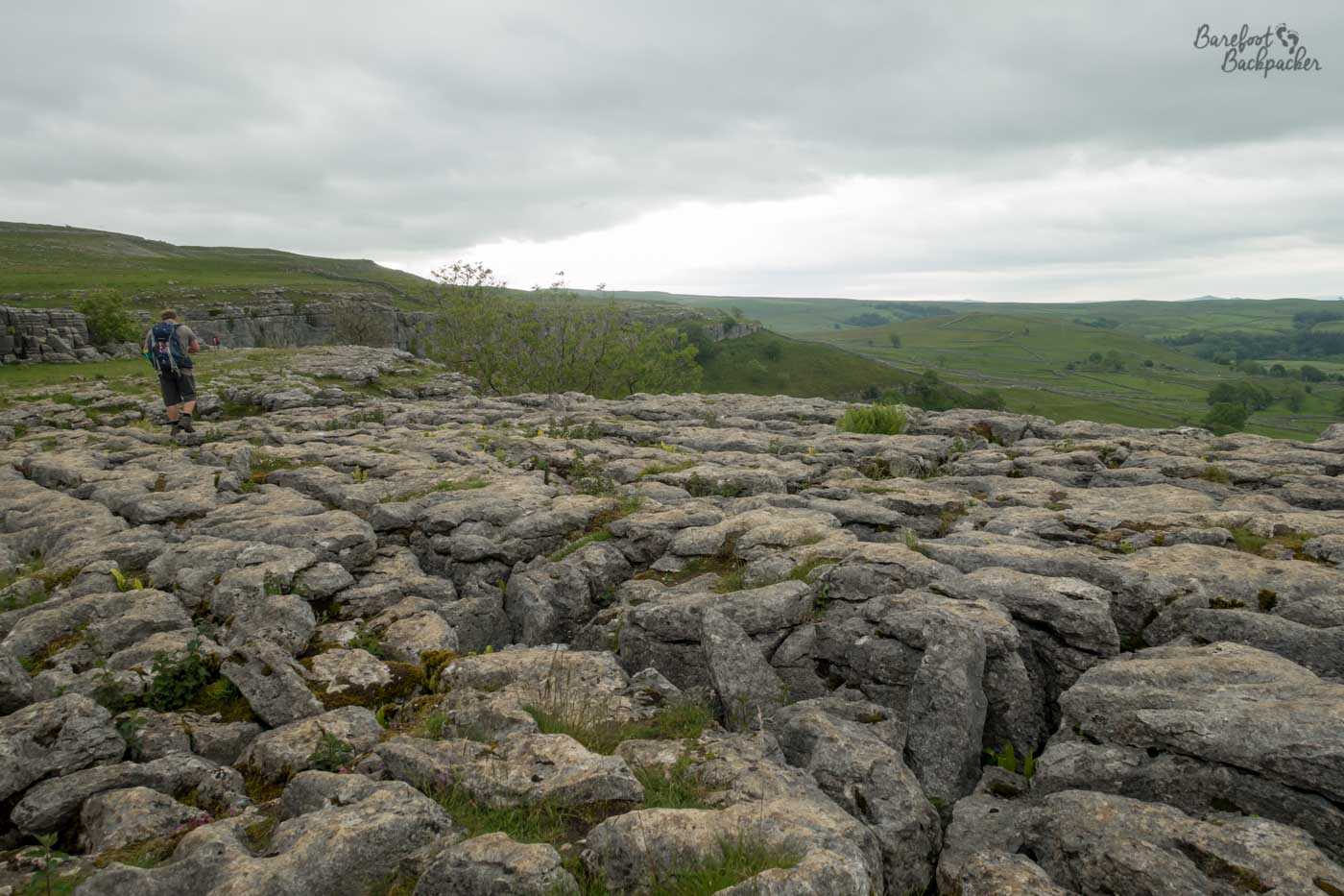 Ragged rocks cover the bottom of shot; they drop sharply off mid-shot and distant, lower, fields can be seen. Someone is walking on the far left of shot over the rocks.