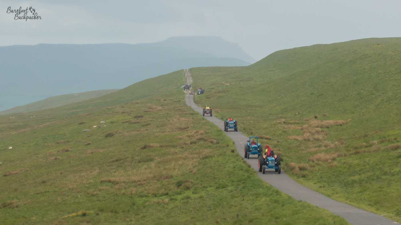 On a country lane running through open moorland, in the distance, are a line of coloured tractors.