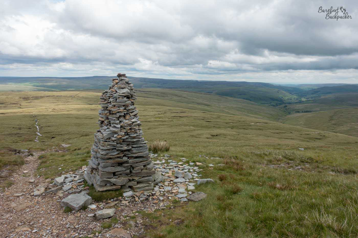 A cairn – a pile of small pebbles/stones made into a small tower. It's on top of a hill. A path descends to the right of shot. On the left the hill rolls downwards steeply and the view is replaced by several other similar hills covered in low-level of grass. The sky is light but full of cloud.
