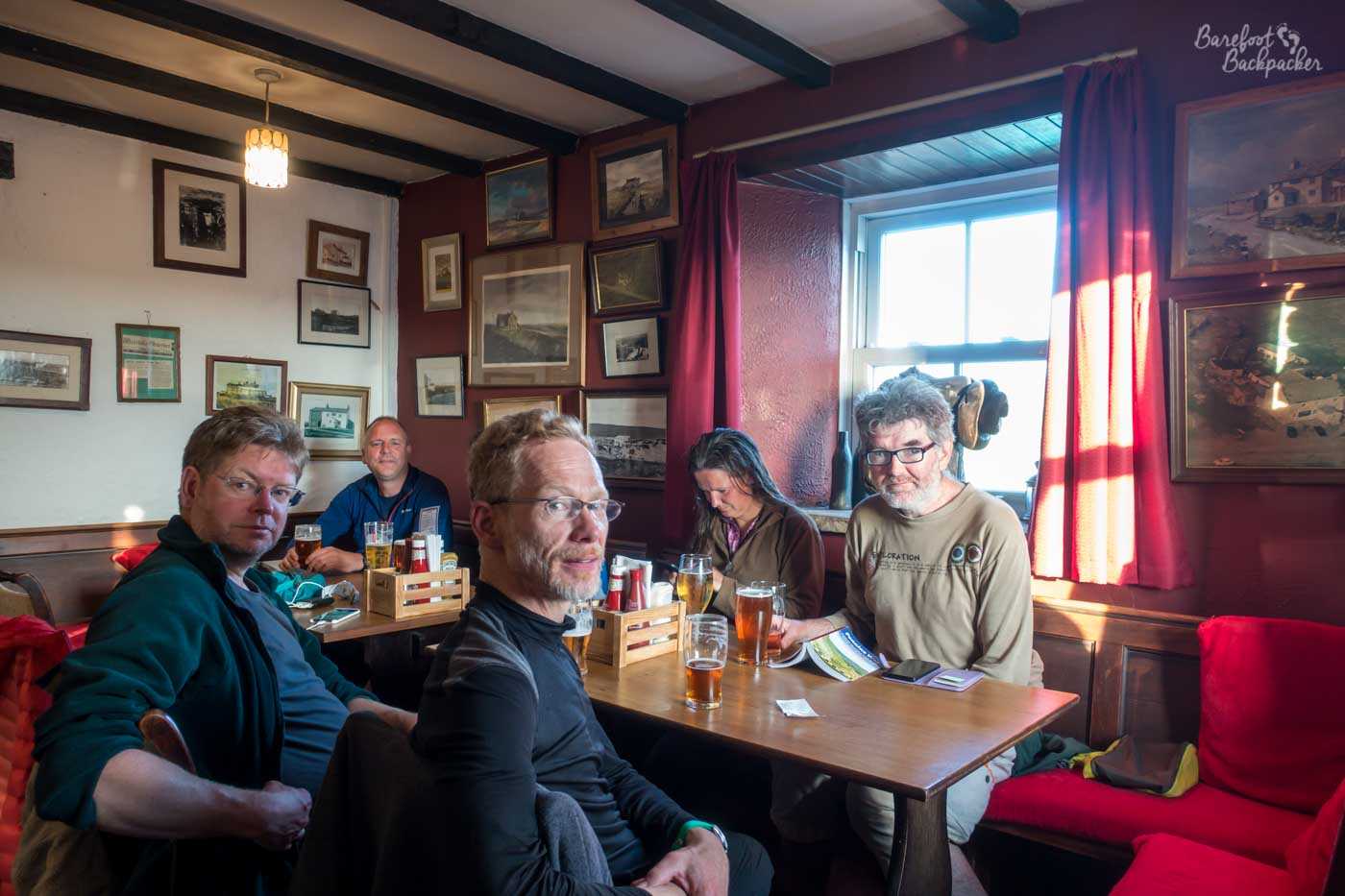 Five people sat around two tables in the pub are facing the camera – well four of them are, one is playing on her phone. The pub walls are covered with old pictures of days gone by. They all have drinks, and there's condiments on the table suggesting food will soon arrive.