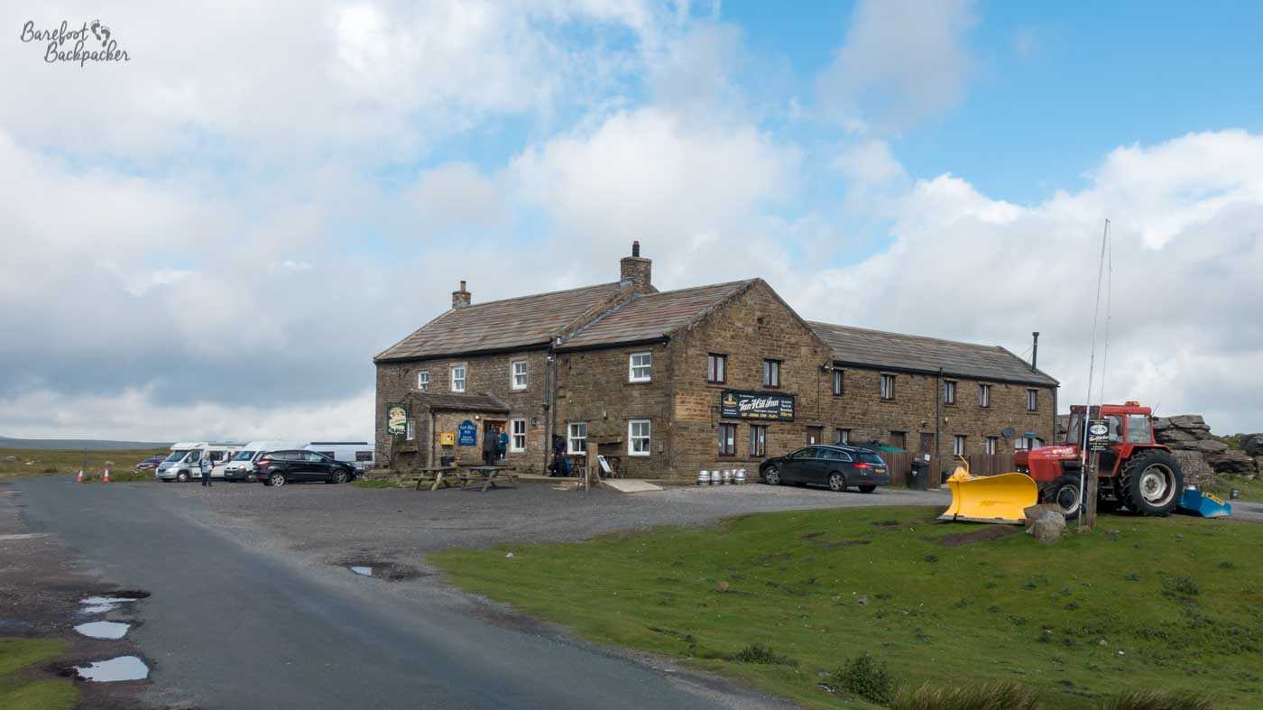 The Tan Hill Inn. It's a large stone building built in what seems to be the middle of nowhere. A road passes by, and there's a couple of cars and a tractor around it, but there's nothing else but empty space all round.