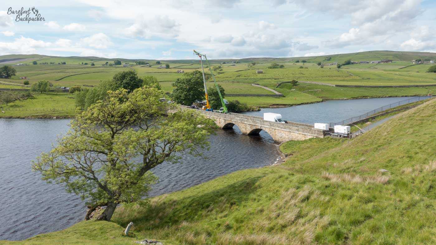 Rolling low grassy hills either side of a reservoir. In the foreground is a tree with branches, hiding some of it (the shot is looking slightly down on it). Across the reservoir is a stone arched bridge. On the bridge are a couple of vans and a huge crane.