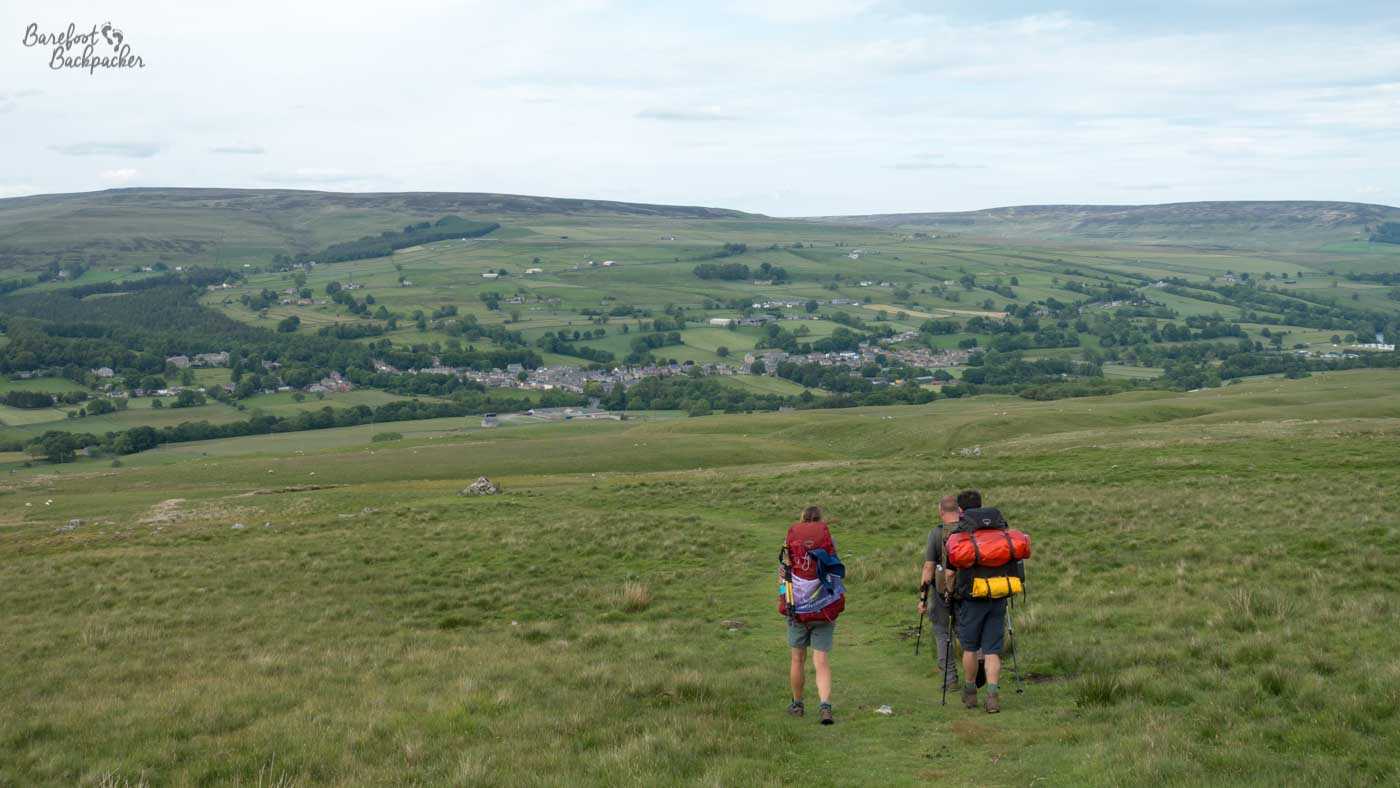 A landscape on both sides of a hidden distant valley. The land slopes gently down towards the middle of shot in both the fore and backgrounds. The background is fileds, some trees, scattered distant buildings. The foreground is entirely grass/shrubs, and three hikers are descending the hill with backpacks and hiking poles.