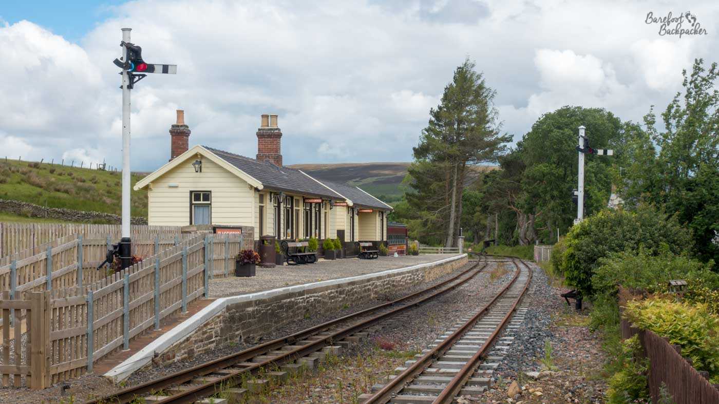 Stone platform. One railway track runs next to it, another runs alongsude that track but with no platform serving it. The building on the platform looks like one of those old Victorian/Edwardian railway station waiting rooms. There's a couple of large poles with signals on them – set to stop. The rest of the environment is hills and trees.