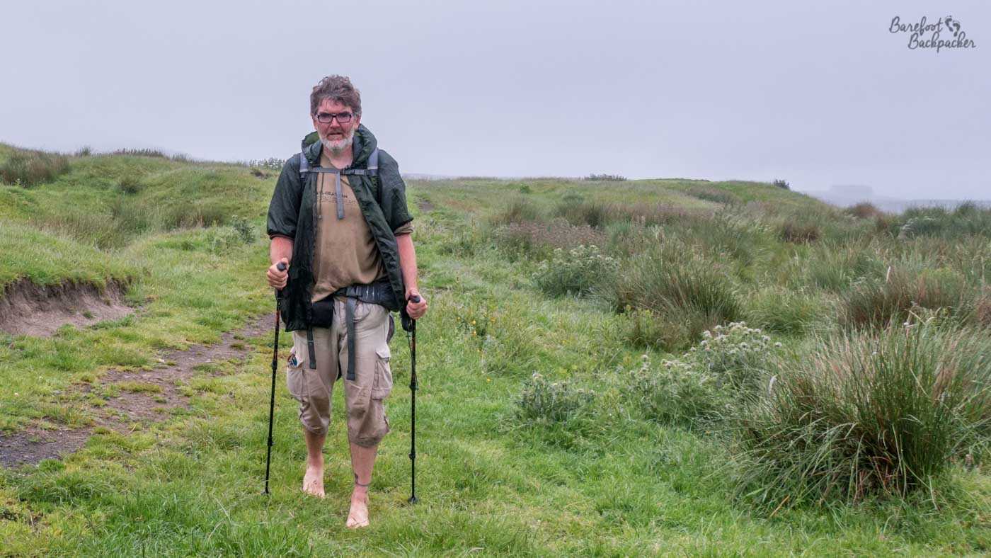 A hiker heads towards the camera. They are holding a hiking pole in each hand. They have a t-shirt, a kagoul, some very long cargo pants, and glasses. They are barefoot. They don't look happy. They give the vibe of being a cross between Gandalf and a hobbit.