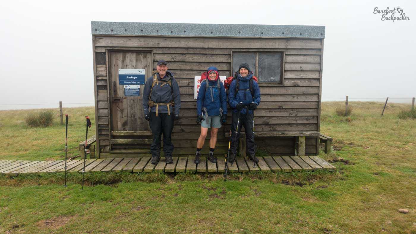 Three hikers stood outside a hut in misty wilderness.