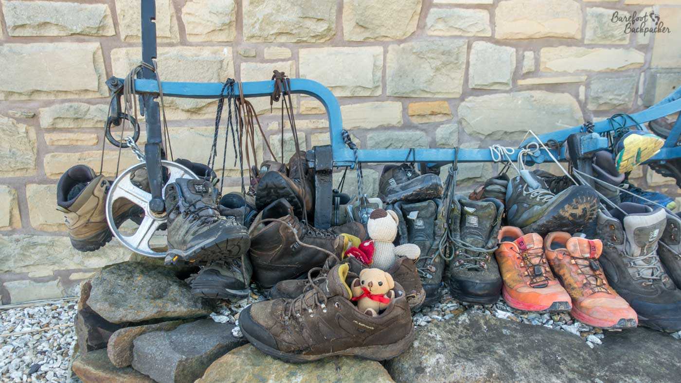 A line of walking boots on a stone wall, mostly with their laces tied to a metal frame beam just above the wall. Two small soft toys are sat in a pair of walking shoes.