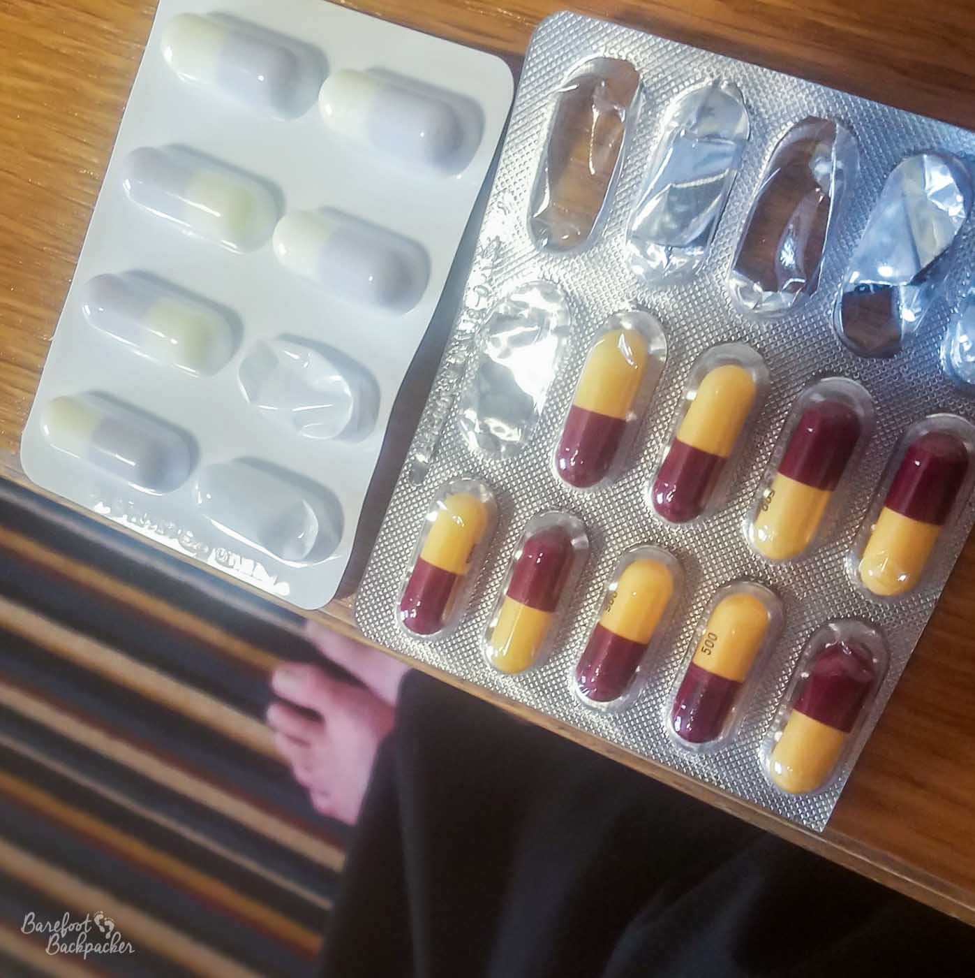 On a table in a hotel room are two packets of pills. The pills are quite big, and each half of the pill is a different colour. Some of the pills have been taken already, evidenced by empty and squashed sections of the pill packet.