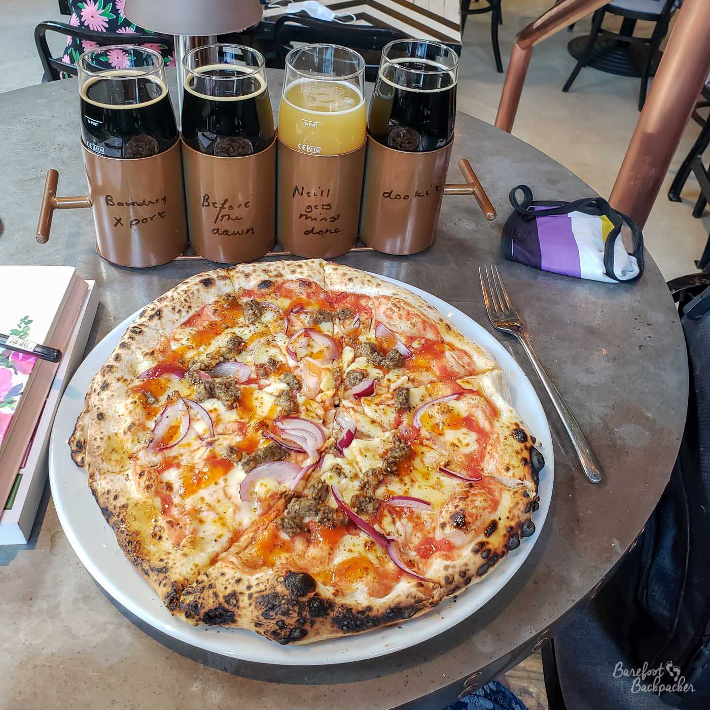 In a pub, on a table. There is a whole circular pizza on a plate, and behind are four small beer glasses in a flight. There are a couple of books just in shot on the left, and a non-binary flag coloured facemask on the right.