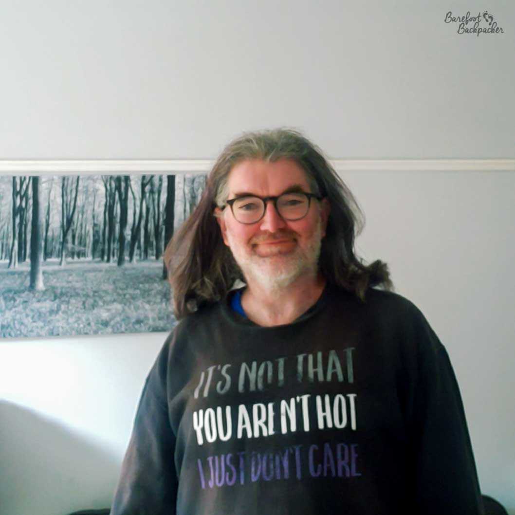 A person is stood in front of a white wall. The image is a head/torso shot. They are smiling at the camera, sort of, and wearing a sweatshirt that says 'It's not that you're not hot, I just don't care', in the asexual pride colours.