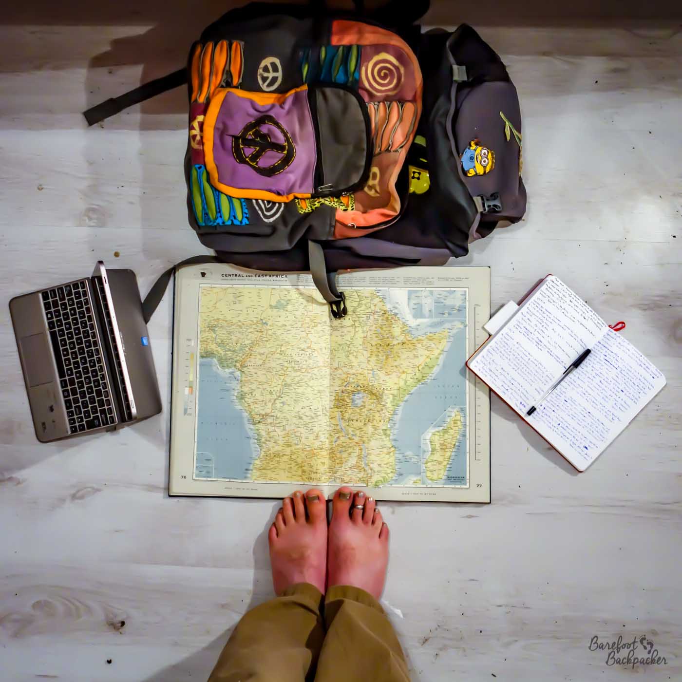On the floor in a bedroom; the floor is lain with laminate slats. In the centre of shot is an old world atlas, open to a page on Central Africa. At the top of it is a fabric daypack, to the left is a tablet computer, to the right is a notebook and pen, and below the atlas are a pair of bare feet.