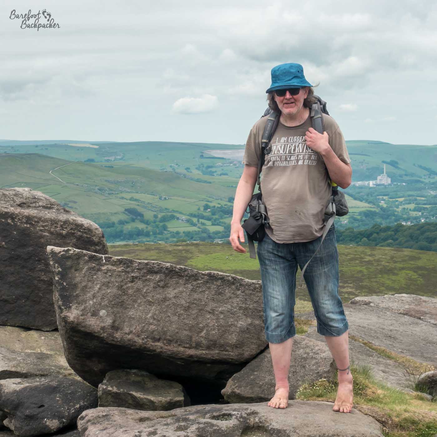 I'm standing on a boulder in the countryside, in front of a long landscape behind with distant hills and bushes. I'm wearing a hat, a t-shirt, knee-length denim shorts, no footwear, and a large backpack.