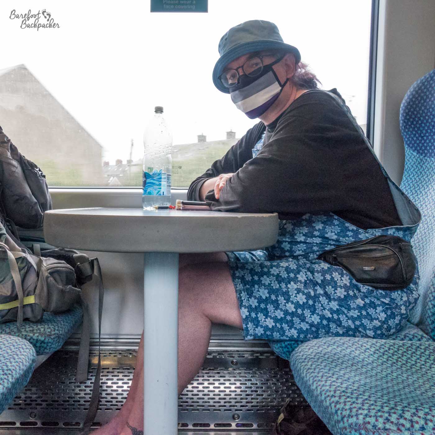Sat on a train, at a table, with a backpack on the opposite seat, I'm in front of a large carriage window looking at the camera. Hat, asexual pride mask, long-sleeved t-shirt, dungaree shorts, and bare feet.