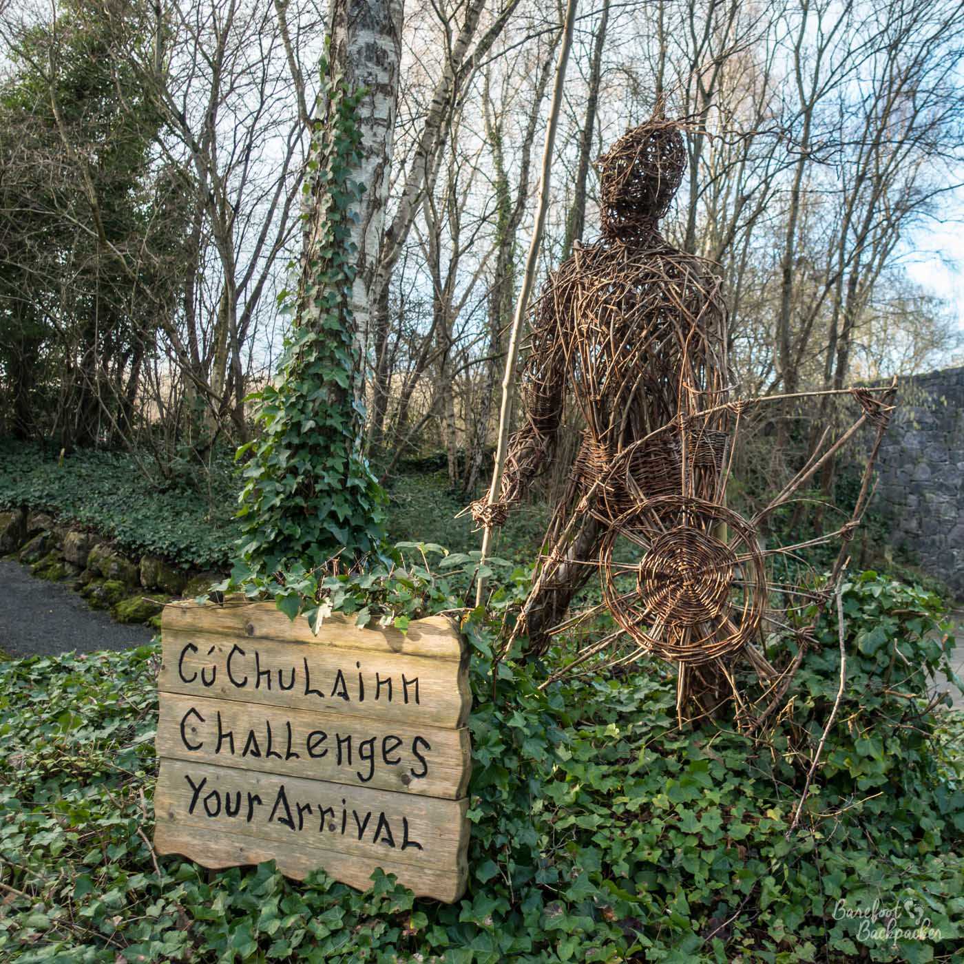 A wicker figure, depicted with a spear in his right hand and a shield in his left, stands in a low-reaching bush in an island surrounded by small roads. In the background are trees. To the figure's side is a large sign; a wooden board on which is written, in stylised Irish script, the words 'Cu-Chulainn challenges your arrival'.