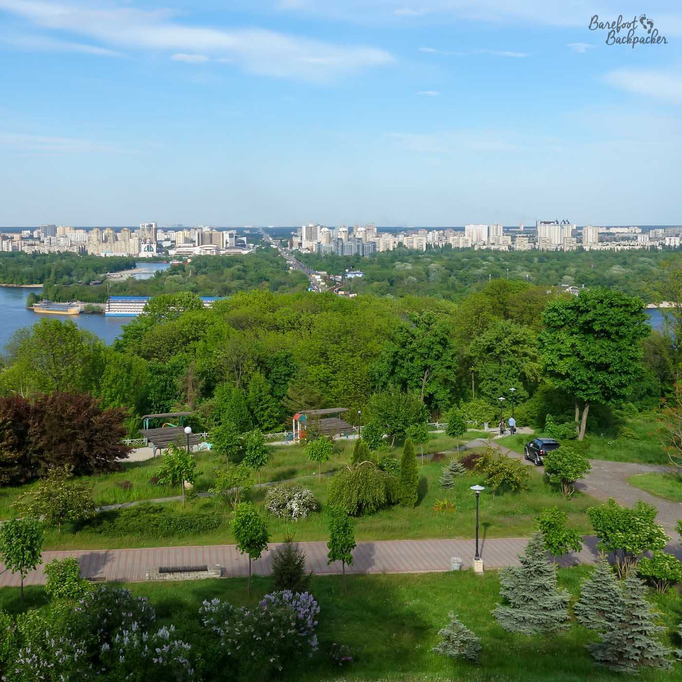From the top of a small hill, there is a view looking out over the landscape beyond. It's mostly trees down below, then a wide river, and in the distance, tower blocks of a different city.
