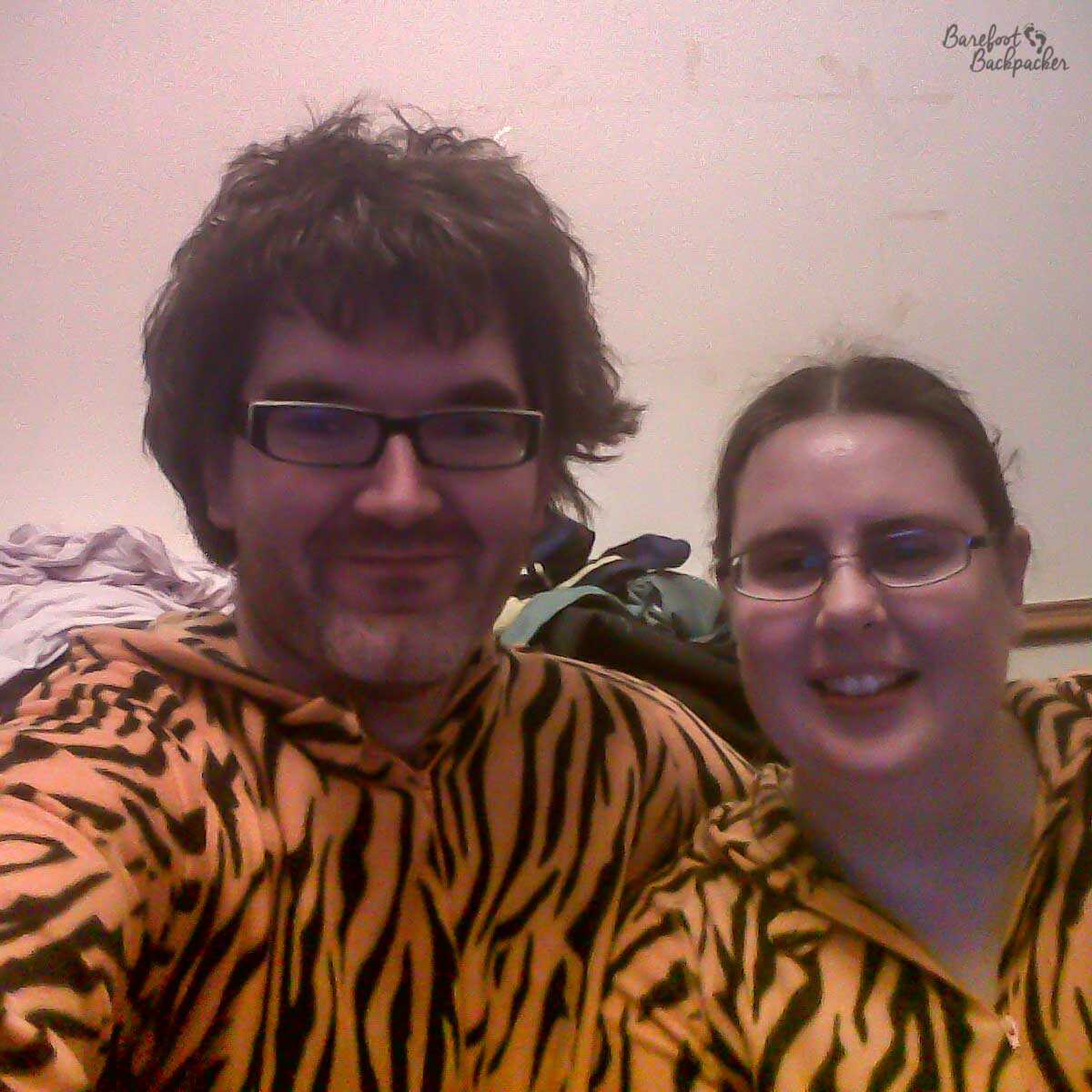 Two people are sitting in front of the camera, in a slightly dark room, wearing matching tiger-striped onesies. One is male, the other female.