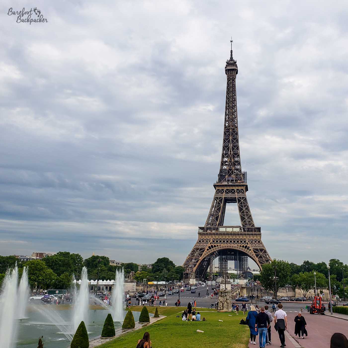 Moody cloudy skies in the background lurk behind an image of the Eiffel Tower, a toer made of a framework of metal, starting at arches and tapering to a point at the top. In front are a busy street, some low trees, and, in the foreground on the left, a couple of small geyser-like water sprays near where some people are sat on grass.