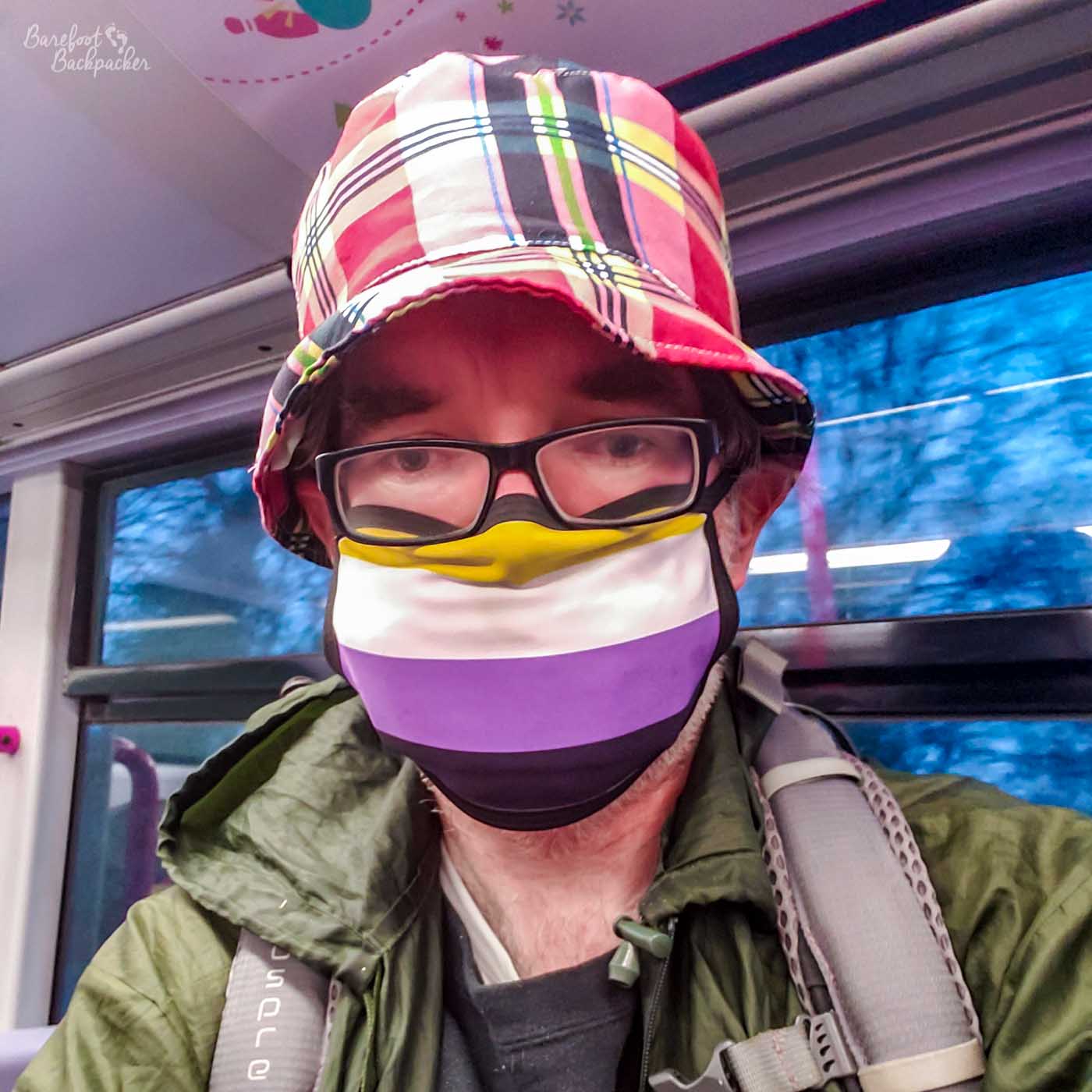 Close-up of a face on public transport wearing a non-binary face-mask, and a tartan-tyle hat. They also have a backpack.