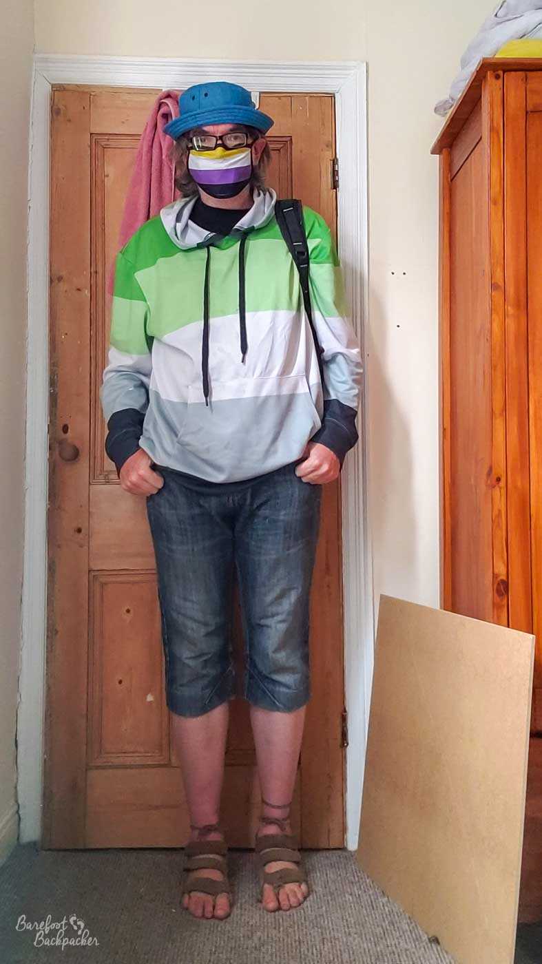 person in front of a door in a room. They're wearing a hat, a hoodie, and jeans that end just below the knee. The hoodie is striped with the colours of the aromantic flag.