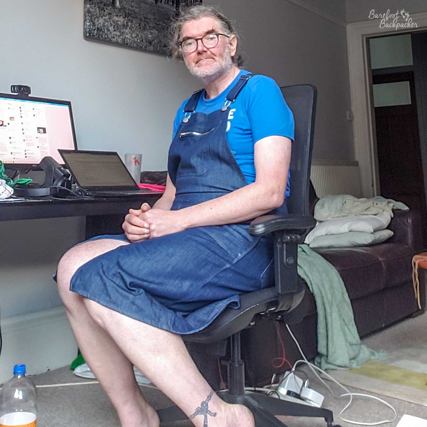 person sat on a chair in a living room, by a computer desk. They're wearing a t-shirt with a denim dungaree dress over it. The dungaree reaches to their knees but there's a dress-like slit in them, revealing more of their leg than dungaree shorts would. They're also barefoot.