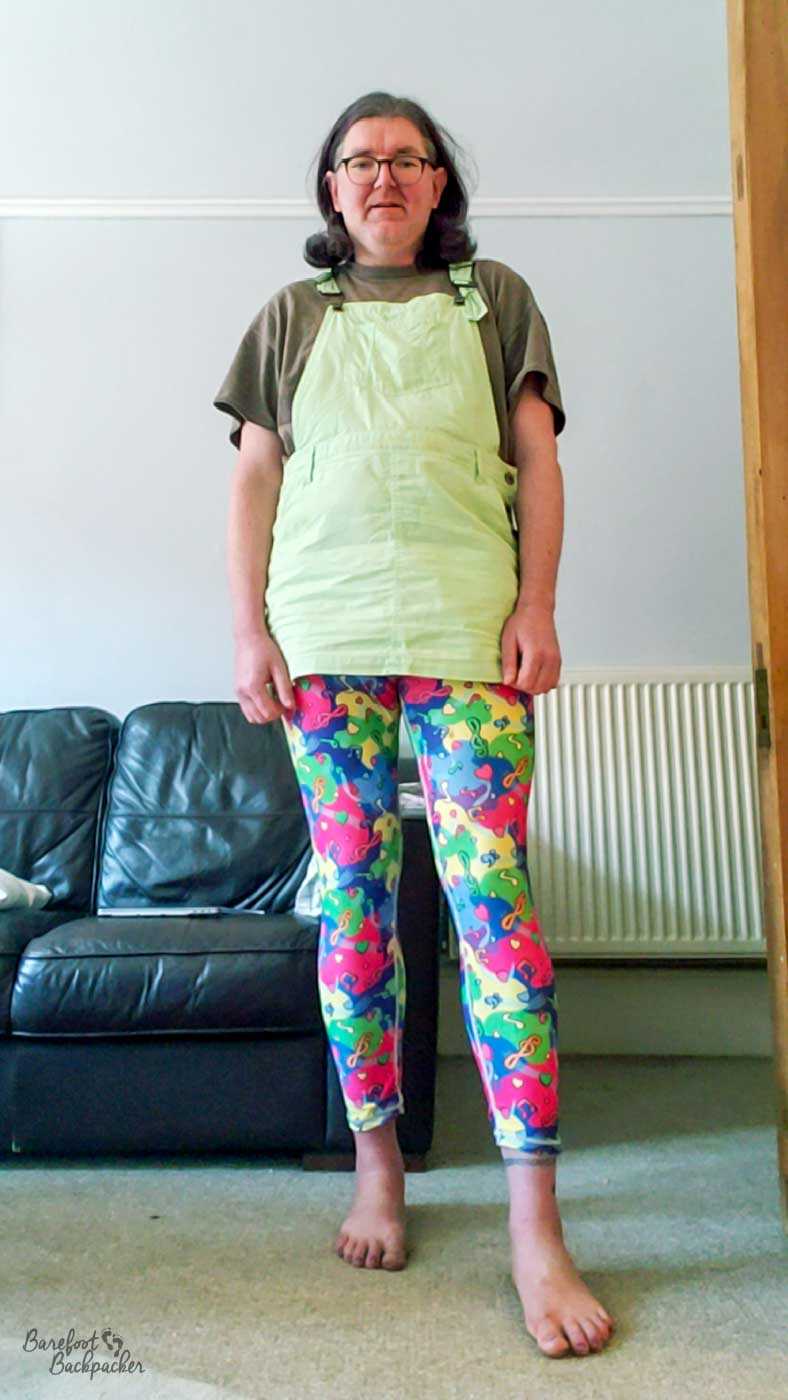 person standing in a room. They're wearing a t-shirt, a dungaree dress that reaches just slightly below their crotch, and some very vibrant and colourful leggings with a music motif. They're also barefoot.