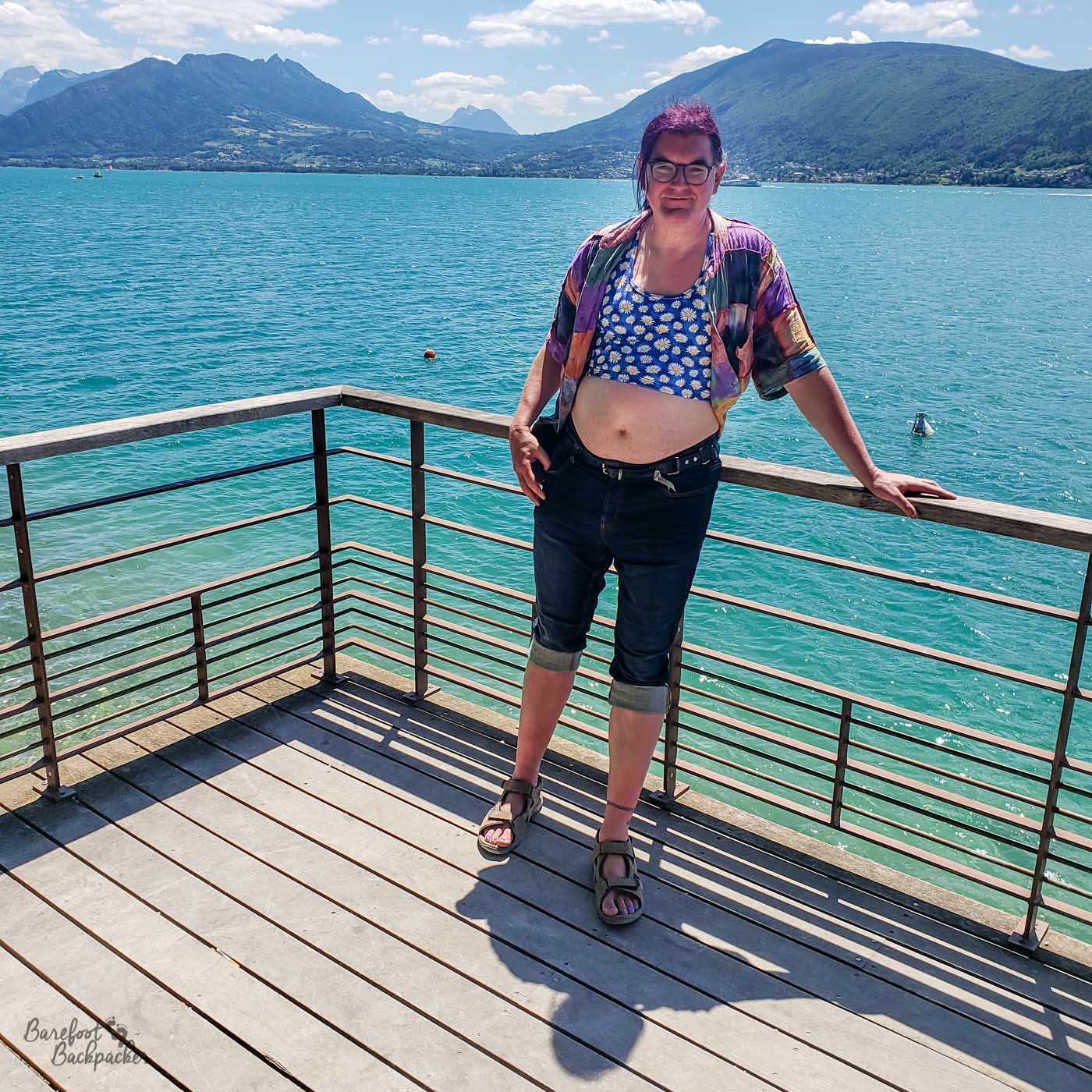 person stands on a boardwalk, with a lake, mountains, and clear skies behind them, They're wearing a patchwork shirt that's fully open, revealing a crop-top with a daisy motif. Their stomach is bare. They're also wearing jeans that are cuffed just below the knee, and sandals.