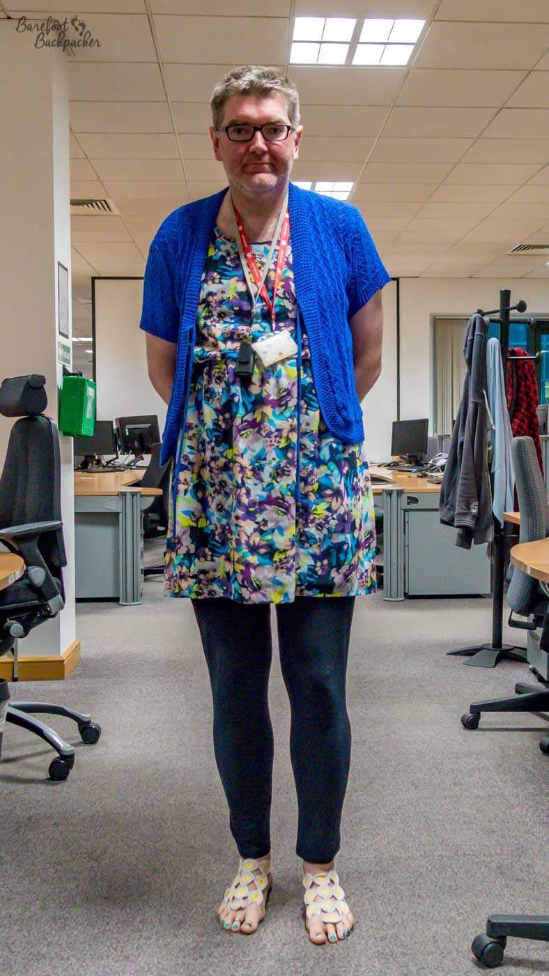 person stands in an open-plan office, around desks and monitors. They're wearing a colourful dress, with a cardigan over the top, black block leggings, and crocheted barefoot sandals.