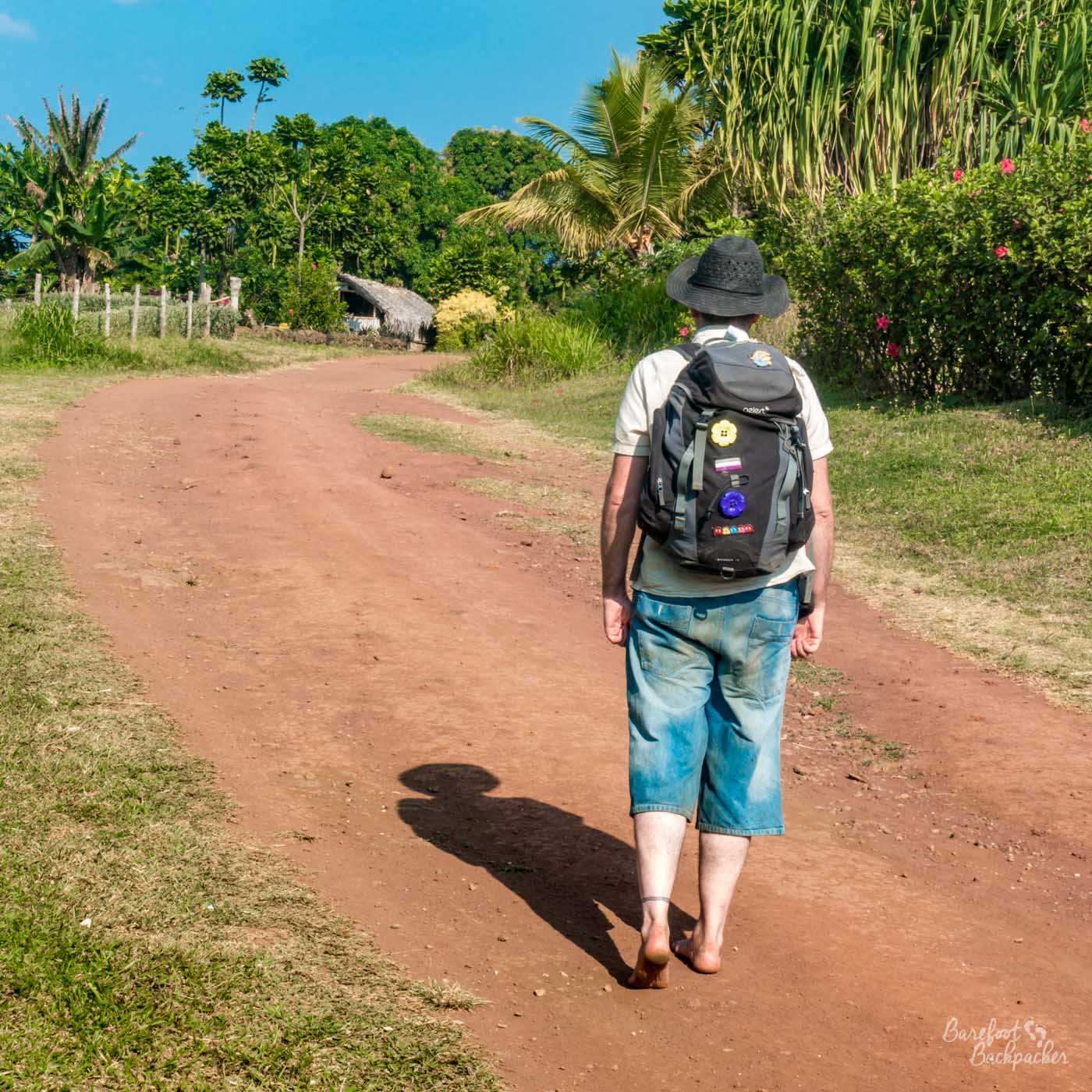 Someone is walking away from the camera down a sandy road. There are trees on one side and a hut in the distance. They are carrying a backpack and are barefoot. There is no-one else in shot.