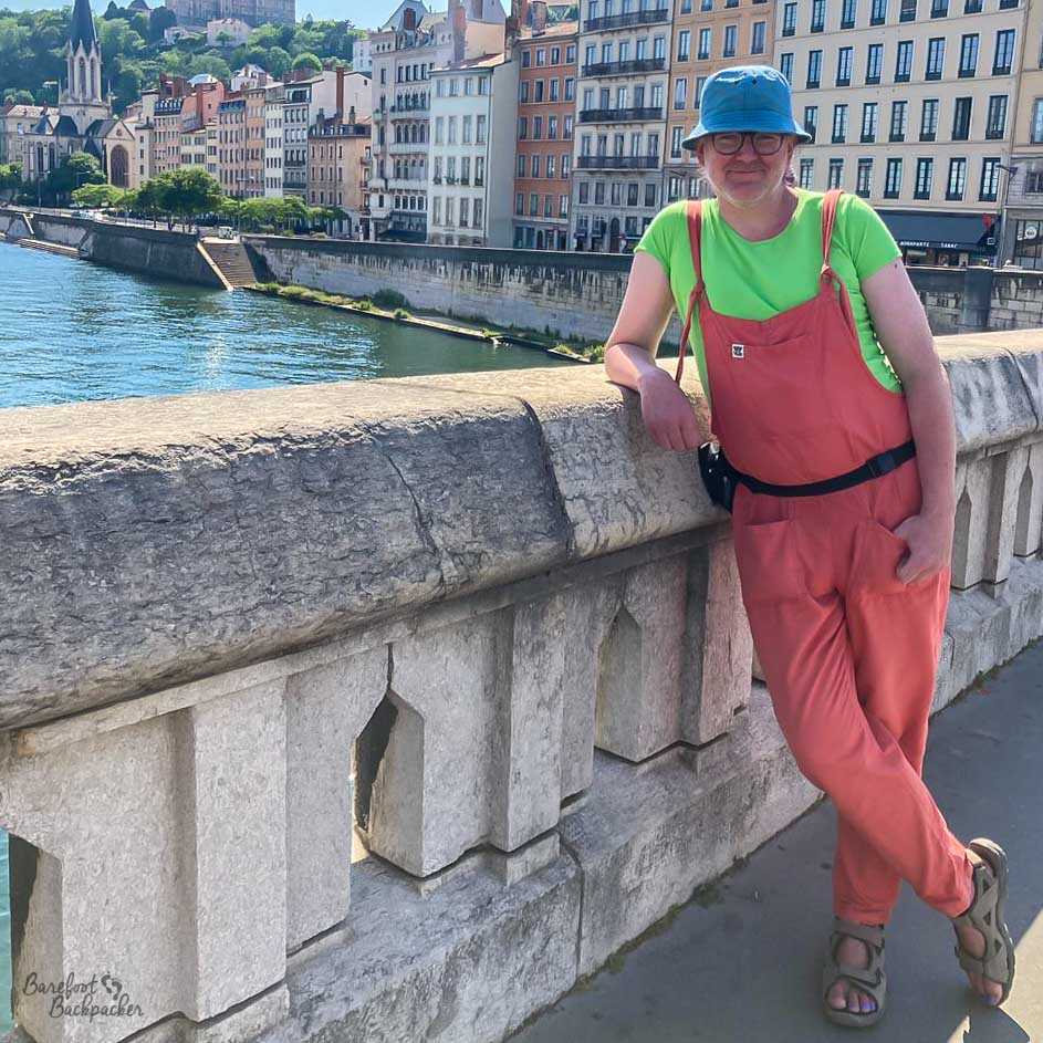 A non-binary person stands on a bridge over a river with buildings in the background. They're wearing long red dungarees and a green t-shirt underneath. The colours involved make them vibe a bit like a watermelon.