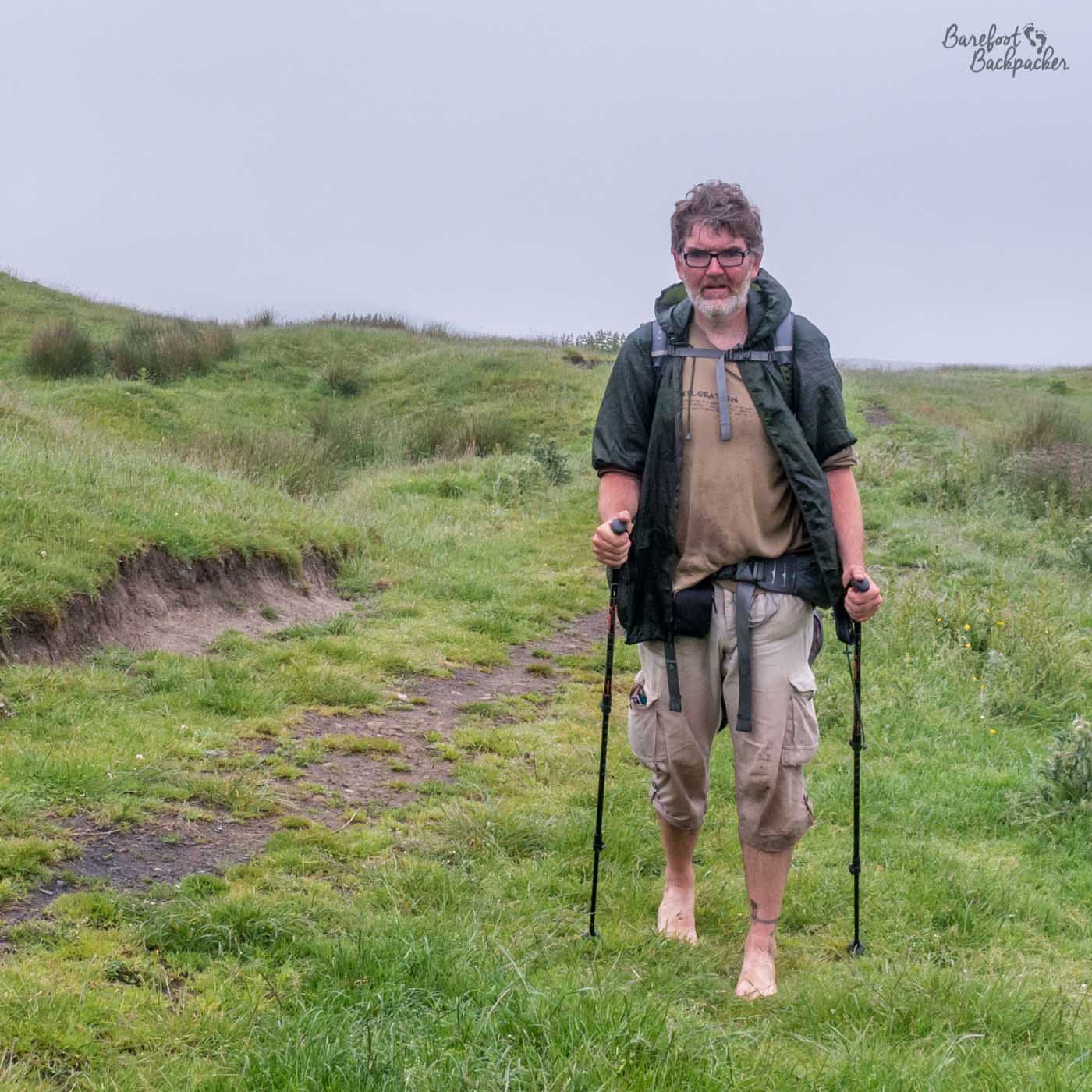Someone is hiking towards the camera through a vague path in the moorlands. They are carrying a large backpack, hiking poles, and are barefoot. They also look very grumpy