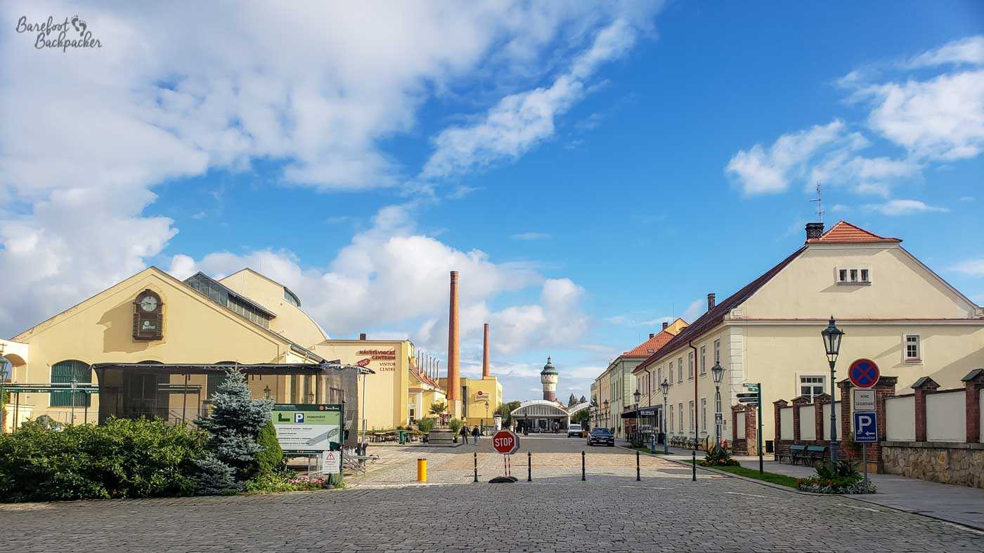 A long cobbled street with buildings on either side. In the distance are some thin tall chimney-like structures and a domed roof covering where the road goes.