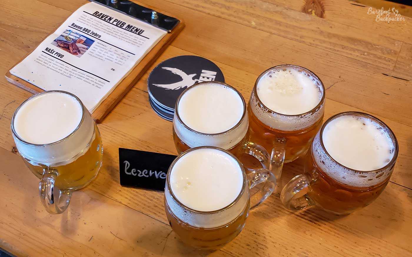Five glasses of beer, with foam heads, are on a table. Also on the table are a clipboard menu and a series of beer mats, both advertising the fact this the Raven Brewery.