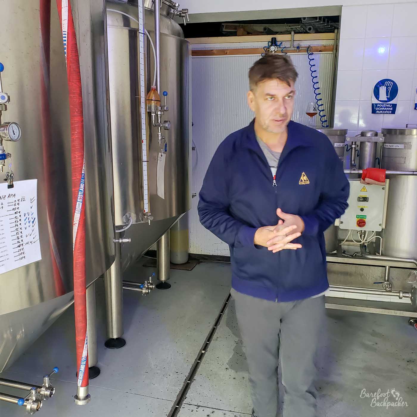 A man, Filip, stands to the side of a series of large metallic beer vats and in front of some industrial equipment.