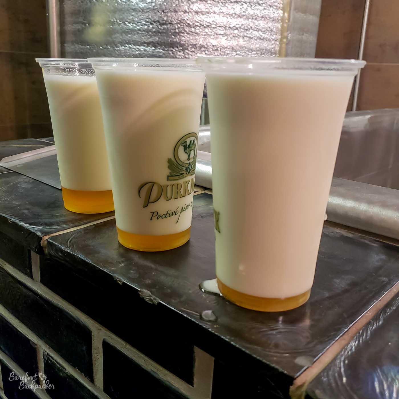 Three large plastic glasses of the Mliko beer style on a tiled shelf.