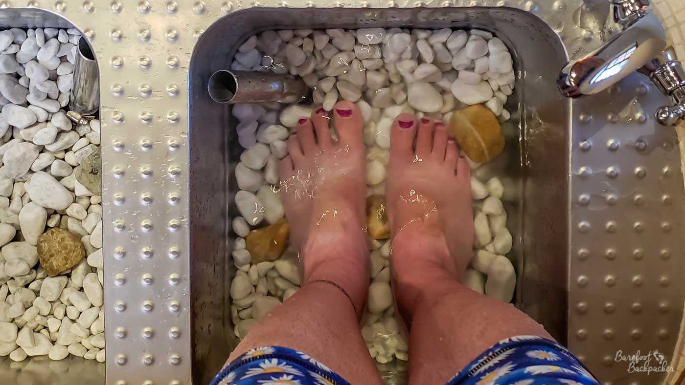 Close-up from above of two bare feet resting in a small metal tank. At the bottom of the tank are stones. The tank is filled with water.