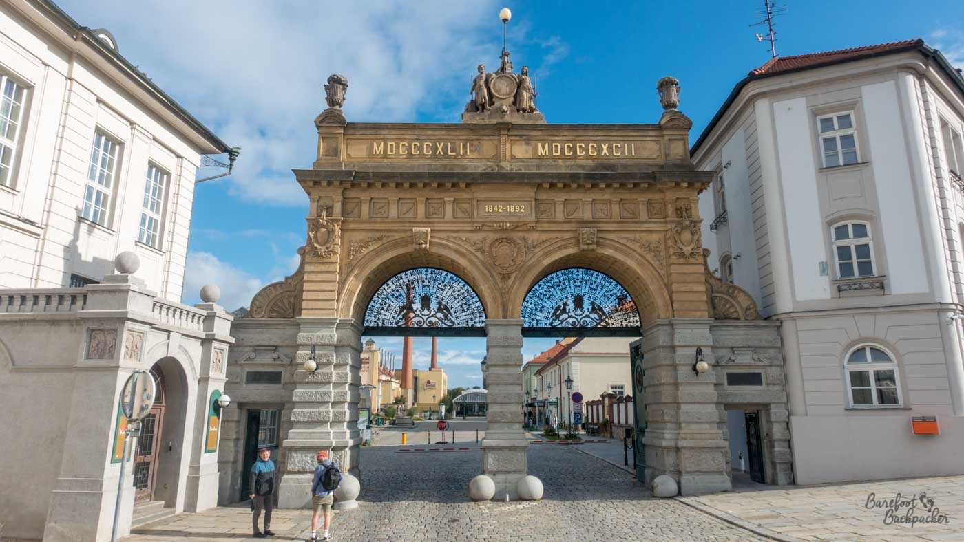 The main gate leading into the Pilsner Urquell Factory. It's a tall stone edifice with two archways covering the road. Above the archways are brass lettering. 1842-1892, in both Arabic and Roman numbers.
