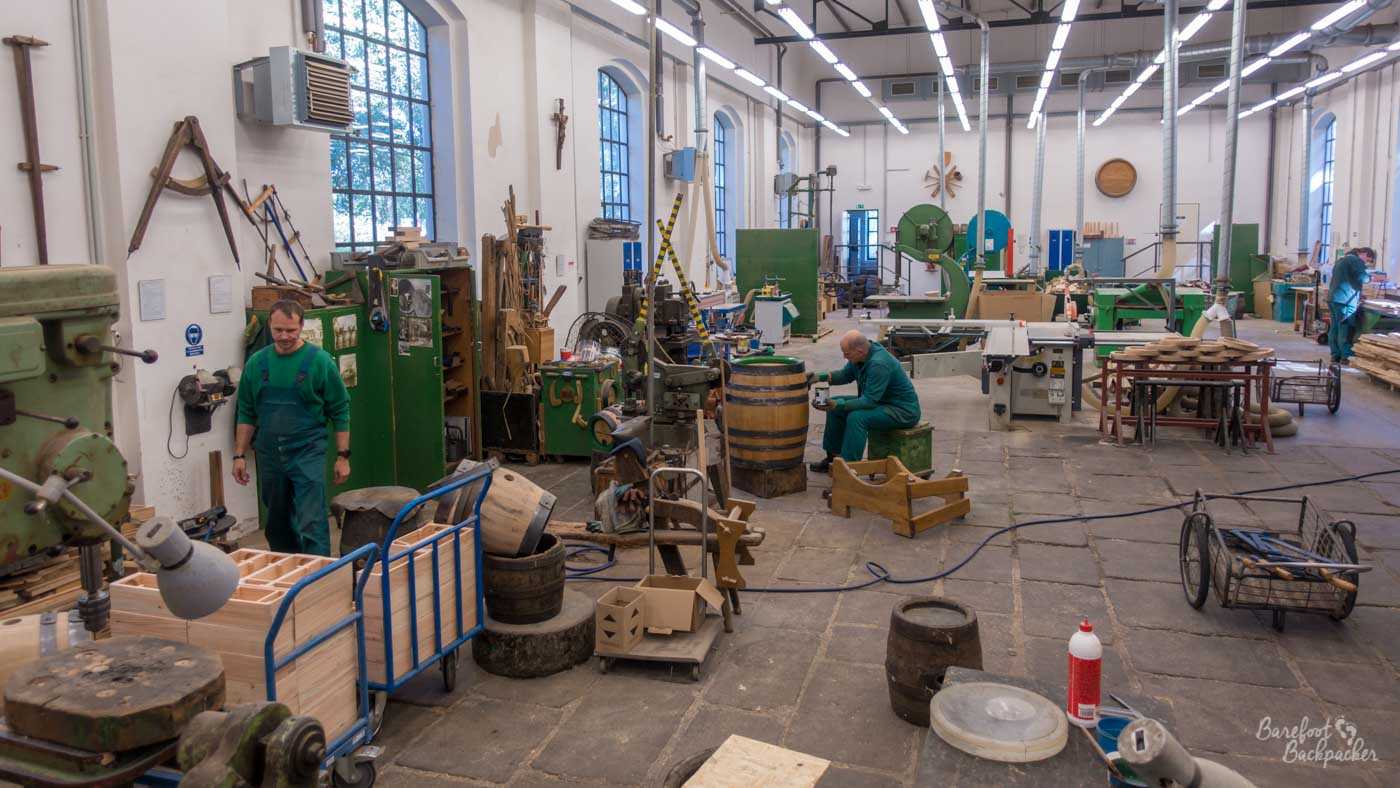 Wide view of a factory floor, with boxes, equipment, tools, and wiring. There's a few people in shot, one of whom is painting a barrel.