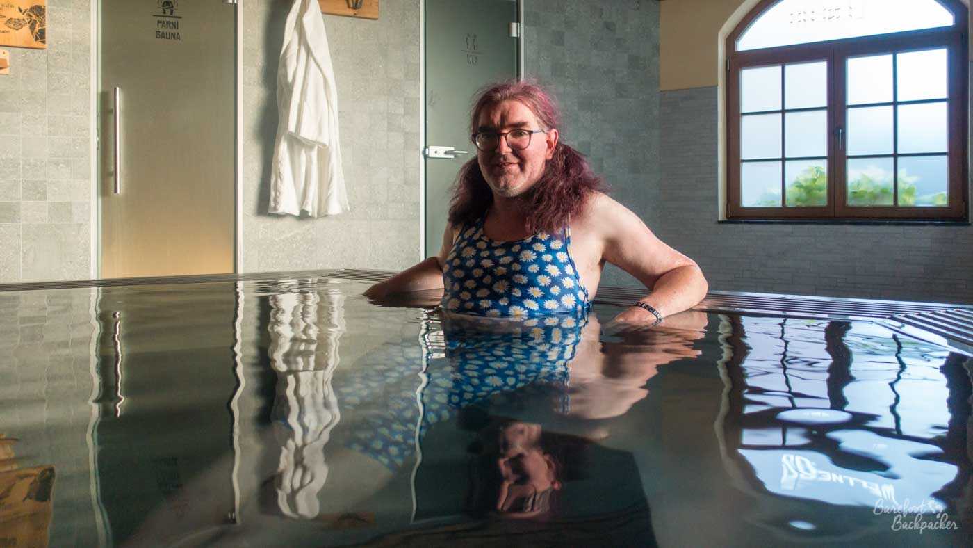 Picture of a person sitting in a hot tub. They're wearing a daisy-motif athletics-style crop-top and capri leggings, and are sat half in the water with legs straight out under the water in front of them. Behind them are the doors to the sauna and shower, a tiled wall, and a frosted window.