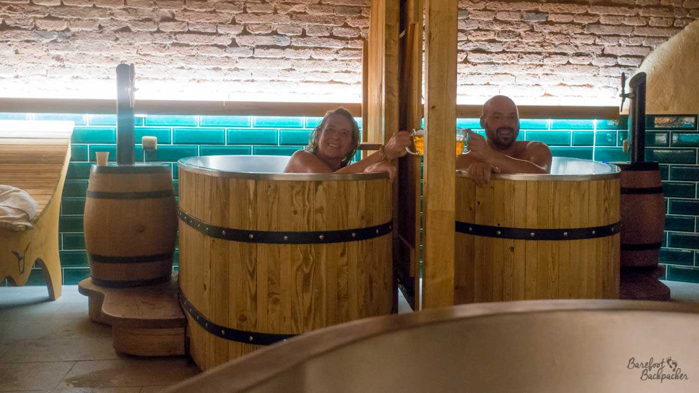Two people are each sat in a separate metal tub, surrounded by wood to make it look like they're in beer casks. They are both staring at the camera while holding up beer glasses to each other in a 'cheers' motion, through a hole in the wooden roof supports.