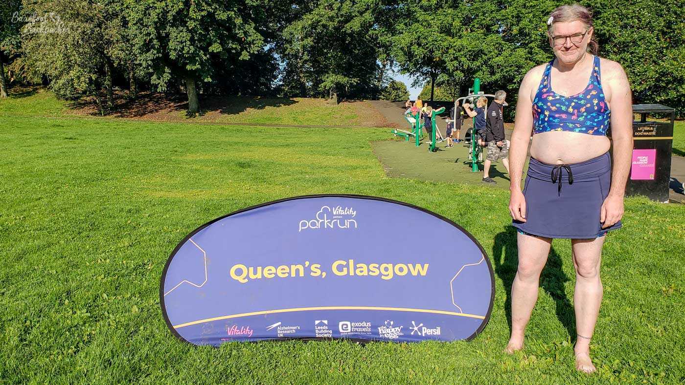 On a grass field with trees and an outside multi-gym area. In the centre of shot, propped up on the grass, is a banner advertising 'Queen's Parkrun Glasgow''. Standing on the right is a person in a crop-top covered with a pattern of coloured cartoonish feet, a plain skort, a daisy in their hair, and, so it seems, bare feet.