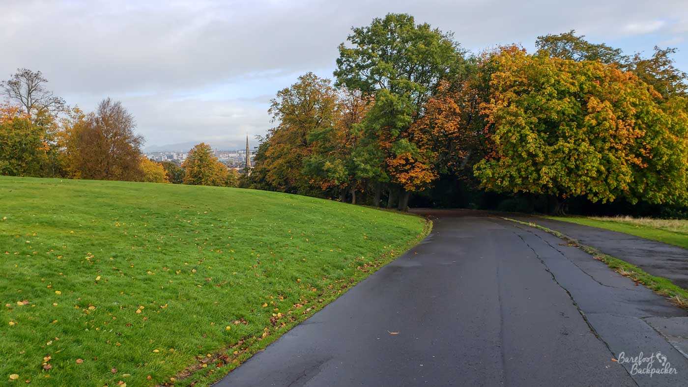 A wide tarmac road goes down a hill. There are trees in the midground. There's a small cityscape in the distance, and grass to the left.