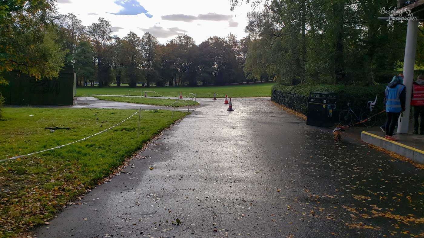 A wide tarmac path. In the centre of it, in the midground, are some traffic cones and police tape – they stay in the centre of the path as it swings left. On the left edge of the path is another line of police tape. This is the finishing funnel for Queen's Parkrun