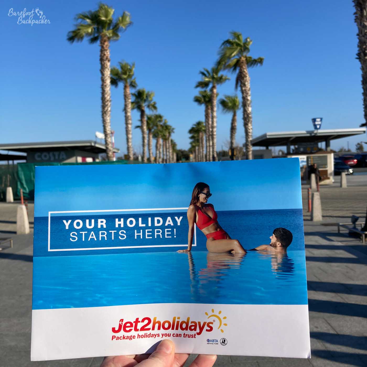 In front of two lines of palm trees, a hand holds up a Jet2 brochure that proclaims 'Your Holiday Starts Here'.